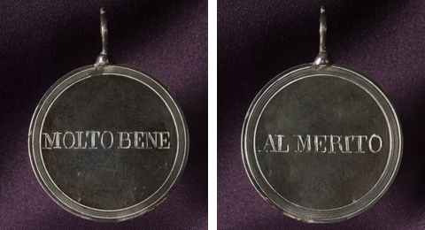 A silver coloured metal disc with a decorative engraved border on both sides. One side of the disc has the words 'MOLTO BENE'. The other side of the disc reads 'Al Merito'. A piece of metal with a hole is attached to the top of the medal to be used for threading on a necklace or similar.