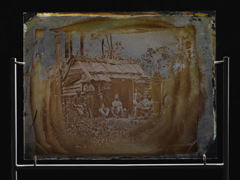 Glass showing seven men outside a bush slab hut. Most of the men are sitting and the man on the far right is only partially visible.