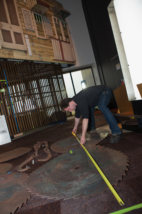 A colour photograph of a man measuring large metal saw blades on the floor.