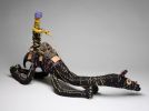 A black and pink model camel and rider made from, raffia, minarri (greybeard) grass, wool and leather. The rider has a yellow and black body, is wearing a purple beanie, and is seated on a brown crocheted saddle which is fastened to the camel with leather straps and leads.