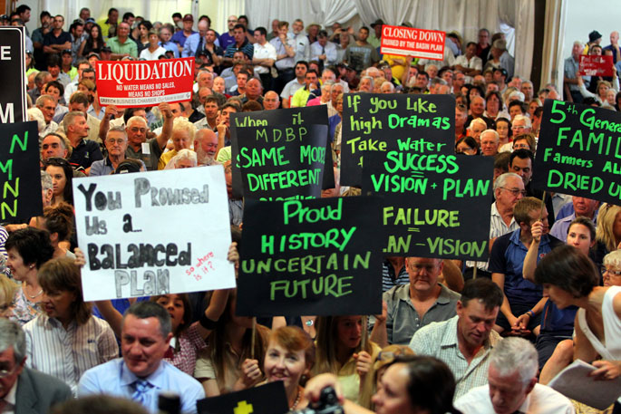 Colour photograph showing a room crowded with people. Some of the people hold protest signs above their heads.