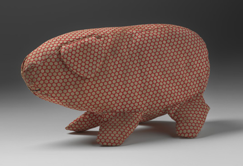 A soft toy in the shape of a pig made from off-white spotted red cotton fabric. The pig has a triangular ear and is missing its tail. A curved mouth is sewn onto the fabric in black thread.