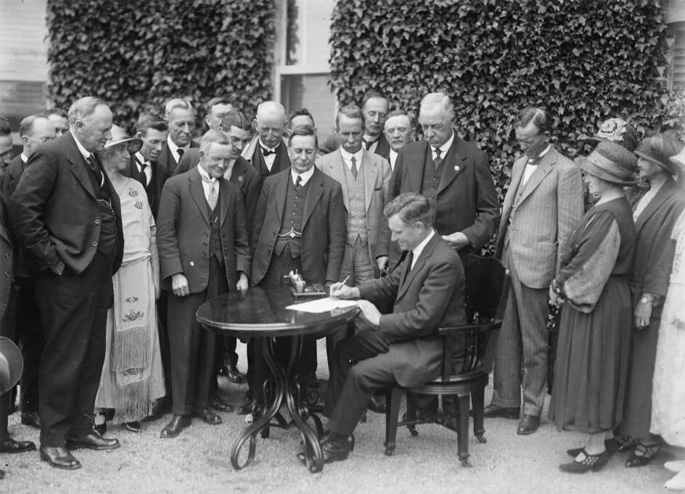 Black and white photo of a group of people watching a seated man at a table signing a document