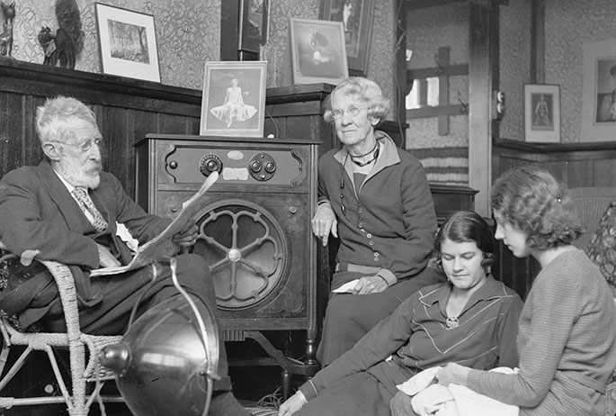 An elderly couple and two young women or girls sit around a radio the size of a bar fridge