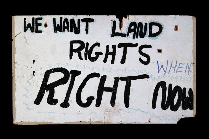 White sign with the words ‘We want land rights. When. Right now’ hand painted in black.