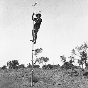 A black and white photo taken in the 1920s of a man who has climbed up to the top of a pole