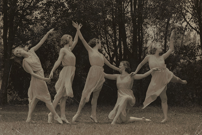 Five ballerinas dressed in white dresses perform on what appears to be grass with trees in the background
