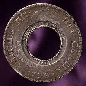 studio photo of silver coin with missing centre. The original Spanish imprinting in Latin is visible around the outside rim, while the words five shillings have been imprinted around the inside rim