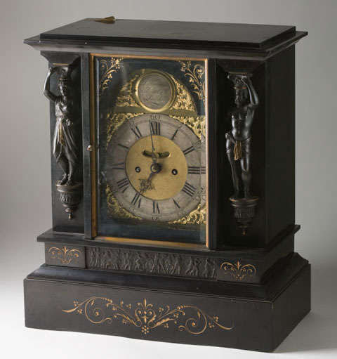 A colour photograph of an ornate clock. The clock has a dark coloured body with decorative features. Small figures can be seen at either side of the clockface. The clockface itself is surrounded by elaborate gold inlay. The base of the clock body has a decorative motif on the side closest to the camera. The entire clock sits on a neutral grey background, suggesting that it has been photographed in a studio setting.