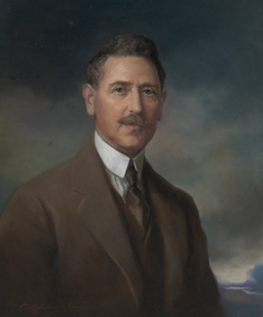 A portrait painting of a man wearing a brown suit and vest, white shirt with a high collar and a dark tie. He has dark neatly combed hair and a moustache. He is visible from the lower chest up. His torso faces toward the right of the painting and he has his head turned toward the viewer. His expression is one of calm and confidence. The background of the portrait is a cloudscape, with clouds in dark tones and a small glimpse of blue sky in the lower right hand corner.