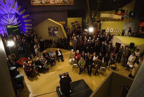 A colour photograph of an exhibition launch in a museum. The photograph has been taken looking down upon the launch, in a 'bird's eye' view. In the foreground of the photograph is a lectern on a small stage. A man stands at the lectern, with his back to the camera. In front of him are two groups of what appear to be official guests for the launch. They all sit on seats. Behind them stand many more people. In the background can be seen what appears to be an exhibition.