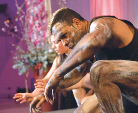 Dancers Ella Havelka and Sani Townson. Townson, at the front of the image, has paint on his arms, legs and face. Both dancers are crouched towards the ground, with arms extended.