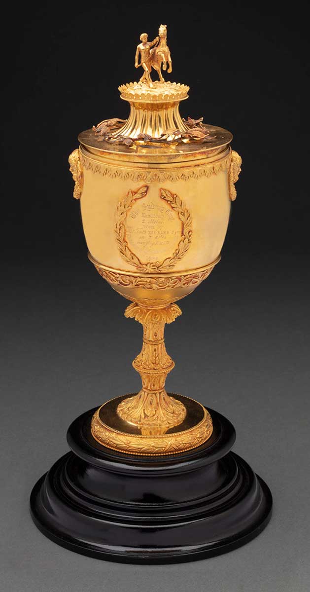 A gold coloured cup on a black circular wooden base. The cup is engraved with the words 'The Sydney Cup / Handicap / 2. Miles / won by / Mr. Taits THE