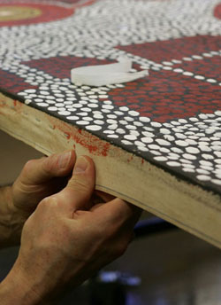 Close-up of two hands extending into the image from the left side. The thumbs are pointing upwards holding onto the side of the canvas of a painting. A small part of the surface is visible and contains white and red dots on a black background.