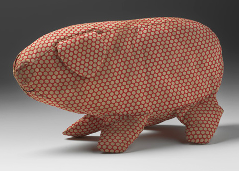 A soft toy in the shape of a pig made from off-white spotted red cotton fabric. The pig has a triangular ear and is missing its tail. A curved mouth is sewn onto the fabric in black thread.