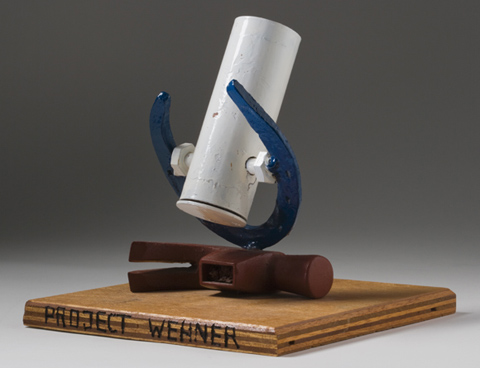 A 'model' telescope with a square plywood base and the words 'PROJECT WEHNER' inscribed at the front. The head of a steel hammer, painted brown, is mounted on the wooden base, and a blue painted horse shoe is welded to the hammer head. The 'telescope' is made of a white painted metal tube, which hangs between the horseshoe.