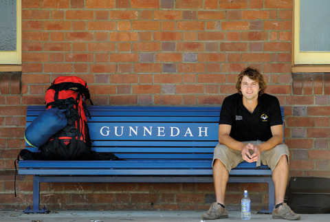 Landscape image showing a man sitting to the far right of a blue, slatted railway bench. 'GUNNEDAH' is painted in white at the centre back of the bench. The man wears a black open-collared T-shirt and khaki shorts and sits with his forearms resting on his thighs. A red and black backpack and a blue sleeping bag sit at the opposite end of the bench. Reddish-coloured bricks form a backdrop.