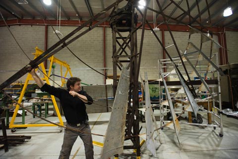 A man extends both arms to turn a large circular section of windmill. Several sections of steel have been attached to the structure, but much of it remains bare.
