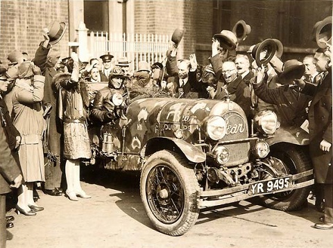 Black and white photo showing a man dressed in leather jacket and cap, sitting in an open-topped car with 'Bean' on the grille. He is surrounded by a group of people who are cheering and raising their hats.