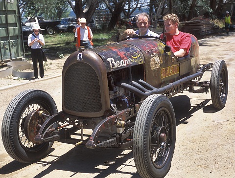 Two men sit in an open-topped car with a small glass panel windscreen.