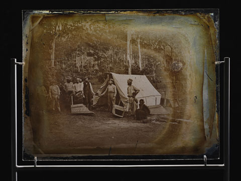 Glass plate showing seven men standing outside a canvas tent, in a bush setting. A child stands to the right of the image, with a woman crouched down beside. Two wooden gold-washing cradles are also visible.