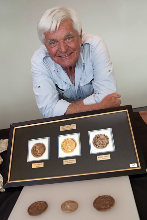 Colour photograph of a man resting with folded arms on a table. In front is a framed set of three circular medals. Another three medals are in the foreground.