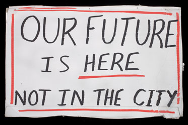 Photograph of a handmade sign on a white piece of card, with a border of red. Black handwritten text reads 'OUR FUTURE IS HERE NOT IN THE CITY'.