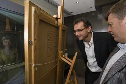 Two men lean in to read words painted on the back of a framed canvas. Part of a portrait of Queen Elizabeth wearing a crown and pale-yellow dress is reflected in a window in front of the painting.