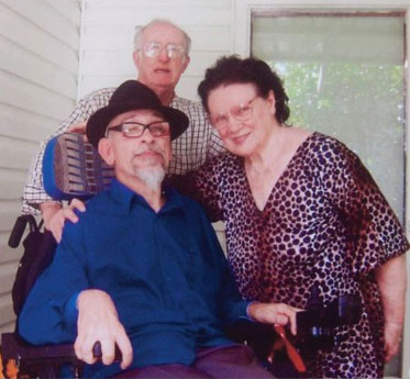 A man sits in a wheelchair. A man standing behind rests his right hand on the seated man's shoulder. A woman leans in to put her right arm around the seated man.