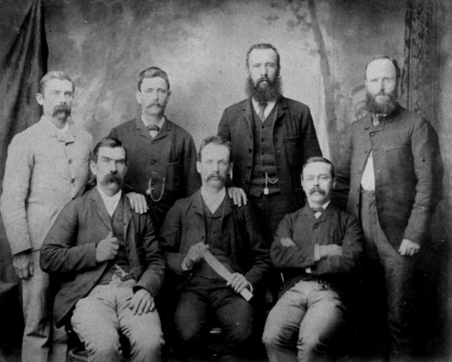 Seven men in a studio group photograph, staring unsmiling at the camera. The name of organisation has been hand-written on the top frame.