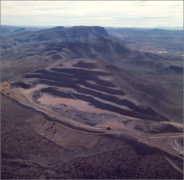 Aerial photo of an open cut mine carved out of the side of a large hill. Massive mining trucks are visible on the perimeter road. Unsullied countryside and mountains in the foreground and background.