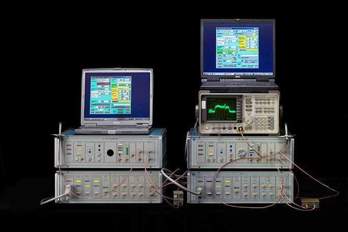 Computer set-up with four grey hardware boxes in two stacks topped by two notebook computers showing data and a spectrum analyser on the right.