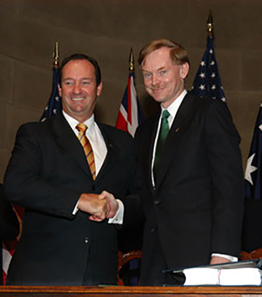Vaile and Zoellick pose for the cameras shaking hands and smiling. Behind them are a series of Australian and US flags along with a man and a woman smiling and clapping