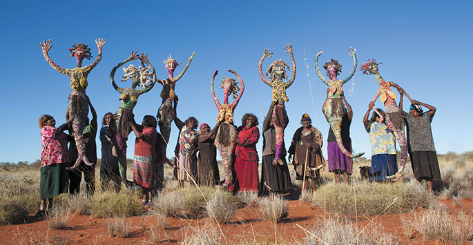 Group of women in a desert holding large grass figures above their heads. <em>Kungkarrangkalnga-ya Parrpakanu — Seven Sisters Are Flying</em> with their makers, the Tjanpi Desert Weavers from Papulankutja, 2015.