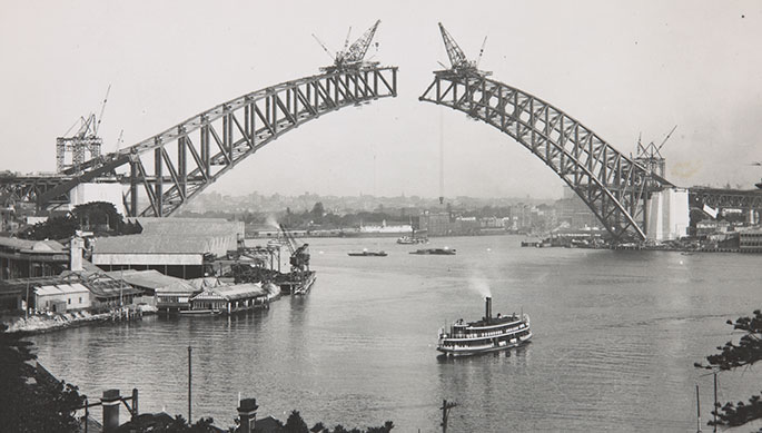 Photo taken from the north shore showing the two sides of the arch under construction. They have yet to meet. Large cranes are perched on top of each end of the spans. A ferry can be seen in the foreground.