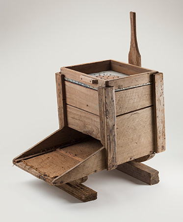 Studio photo of a simple contraption made of timber. It appears to be essentially a box with a tray extending from one side, and a handle from the other. On the top surface of the box is a metal sheet punctuated with holes.