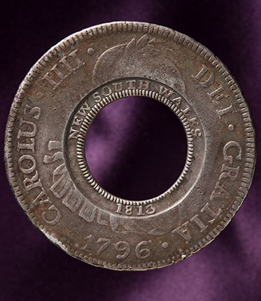 studio photo of silver coin with missing centre. The original Spanish imprinting in Latin is visible around the outside rim, while the words five shillings have been imprinted around the inside rim