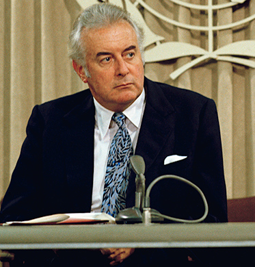 Colour photo of middle-aged man in suit sitting behind desk with UN symbol in background.