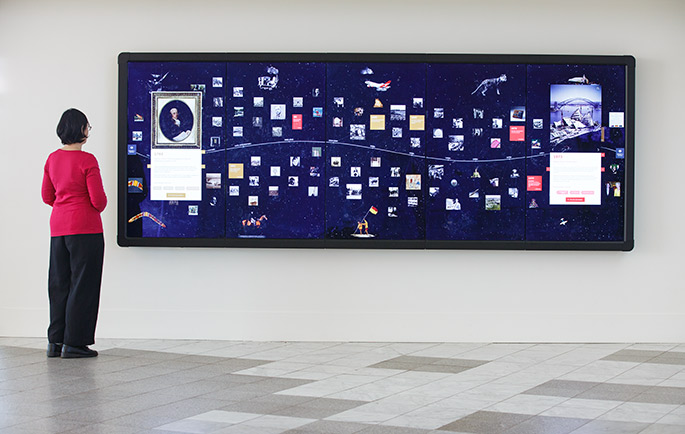 a long digital touch screen mounted on a wall. A woman dressed in red top and dark trousers is facing the screen.