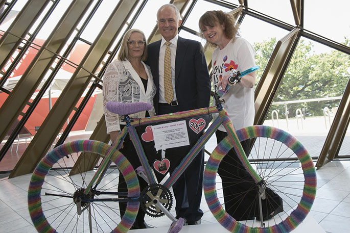 Two women and a man standing behind a bicycle covered with rainbow-coloured yarn.