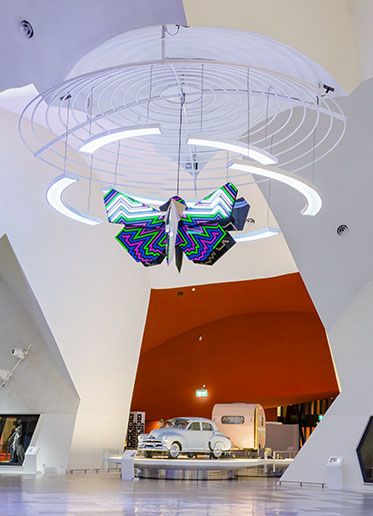 A vast atrium with a brightly-coloured sculpture suspended from the ceiling and a Holden car, with pink caravan, at the centre of frame.