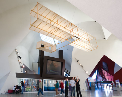 A photograph showing a wood and fabric biplane glider suspended from the ceiling of a museum. The glider has a wooden frame and a box-style tailplane. It is hung with its front end facing the right of the photograph. Underneath the glider are three people, looking up at it. One person has his arms raised above his head pointing at something on the glider. Other people can be seen in the background of the photograph. The ceiling of the photograph has distinctive curved sections that create dramatic angles and forms.
