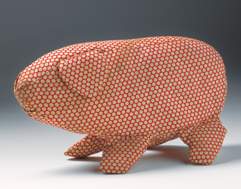 A soft toy in the shape of a pig made from off-white spotted red cotton fabric. The pig has a triangular ear and is missing its tail. A curved mouth is sewn onto the fabric in black thread.