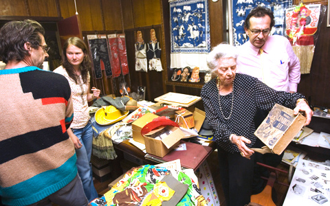An image reminiscent of a dress-ups room. In the front left of the image are two people – a man with his back to the camera, a woman facing the camera but looking to the side. In front of them are tables piled with a variety of boxes, hats, drawing, capes, cloth and other objects. In the background smocks and cut outs of people dressed in costumes are hanging on the wall. On the right side of the image a woman and man are facing the camera and looking at a box that says 'Conductor Set' on the front.