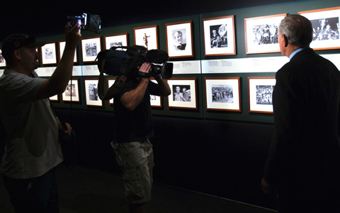 This image of one wall of the exhibition features two parallel rows of framed black and white photographs that stretch across the image horizontally from edge to edge. On the right side of the image Reg Gasnier has his back to the camera and is facing the photographs. A TV camera man faces him with a camera on his shoulder. Next to him another man holds a portable light pointing towards Reg.