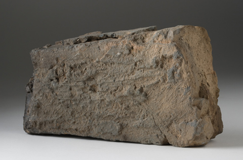 A roughly rectangular brick, greyish brown in colour, with irregular substances on the surface.