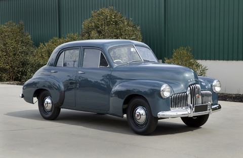 Four door, blue-gunmetal grey sedan with chrome-plated radiator grille, bumper bars, and hub caps. The red entryway to the Museum forms a backdrop.