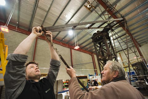 Two men stand to the right of a large windmill frame. The man on the left extends his arms above his head to join two pieces of metal. The man on the right holds one of the pieces in place.