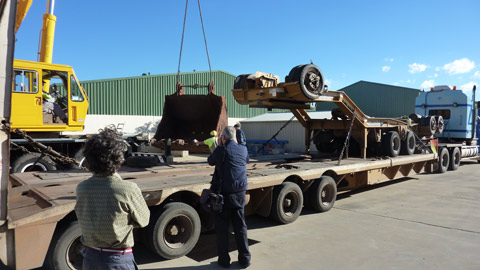 A view of a large mining excavator bucket being lifted off a semi-trailer. The semi-trailer faces diagonally left to right in the image, with its rear at the left-hand side of the image. The bucket has been lifted off the trailer by a large yellow crane, which is just visible at the left-hand side of the image. The bucket hangs just above the ground in front of the crane. Two men are visible near the bucket, possibly guiding it as it's lowered to the ground. Two other men stand in the left foreground of the image, with their backs to the camera. In the background can be seen part of two large green industrial sheds. Above the sheds part of the sky is visible. In the right far distance can be seen a few clouds.