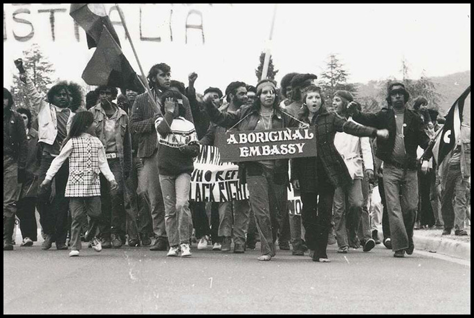 Aboriginal people march, holding flags and banners. A woman in the front has a placard around her neck reading ‘Aboriginal Embassy’